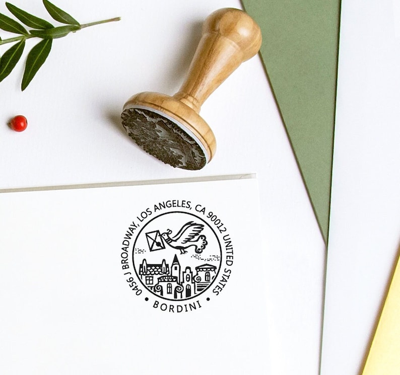 May include: A wooden stamp with a round design featuring a bird carrying an envelope, buildings, and the text "BROADWAY, LOS ANGELES, CA 90012, UNITED STATES, BORDINI". The stamp is on a white surface with green and yellow paper.