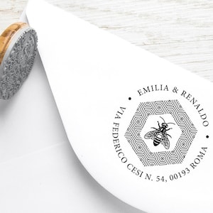 May include: A black and white round rubber stamp with a bee and honeycomb design. The text reads "Emilia & Renaldo, Via Federico Cesi N. 54, 00193 Roma."