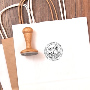 May include: A wooden stamp with a round base and a handle rests on a white paper shopping bag. The stamp has a circular design with text and an illustration of a bird carrying a letter. A second brown paper bag is visible.