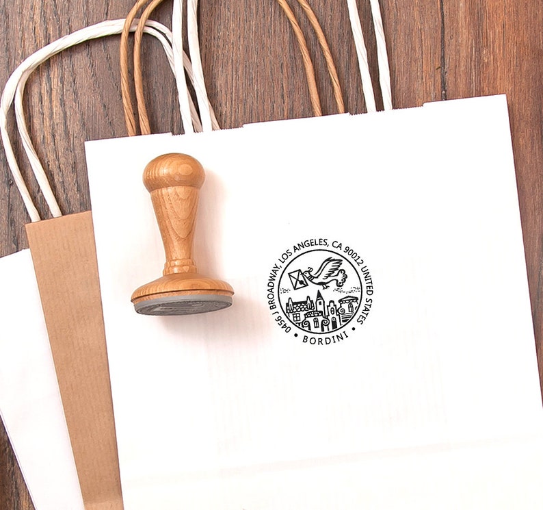 May include: A wooden stamp with a round base and a handle rests on a white paper shopping bag. The stamp has a circular design with text and an illustration of a bird carrying a letter. A second brown paper bag is visible.