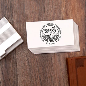 May include: A stack of white business cards with a black circular design featuring a flying bird carrying an envelope, buildings, and the text "BORDINI". The cards are on a wooden surface, next to a silver card holder.