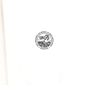 May include: A circular black and white stamp with the text "Broadway Los Angeles, CA 90012, United States" and the name "Bordini". The stamp features a flying bird carrying a letter and a depiction of buildings.