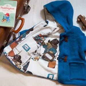 Peut inclure: Un duffle-coat bleu à capuche avec des fermetures à bascule, orné d'une doublure sur le thème de Paddington Bear. Une valise vintage et un exemplaire du livre "Paddington" sont à côté du manteau. Une petite voiture en bois est également présente.