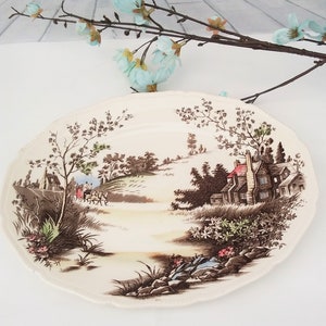 May include: A vintage oval-shaped ceramic plate with a brown and green country scene. The plate features a house with a red roof, a horse-drawn carriage, and trees.