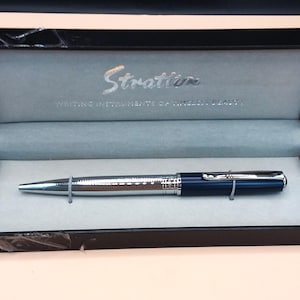May include: A blue and silver pen in a black presentation box. The pen has a silver body with a blue cap and accents. The box is lined with light blue fabric and has the word "Stratton" printed on the inside.