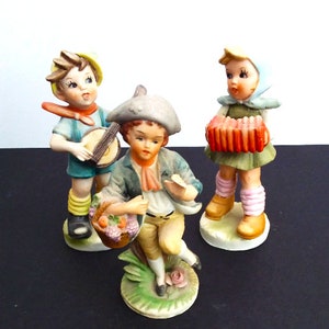 May include: Three porcelain figurines of children playing music. A boy in a blue shirt and yellow hat plays a drum, a girl in a green dress and blue hat plays an accordion, and a boy in a blue jacket and brown hat plays a flute.