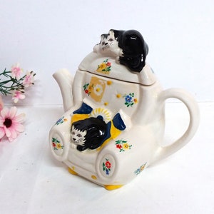 Op de afbeelding: Een witte theepot van keramiek in de vorm van een fauteuil met een zwarte kat die op de armleuning zit en een andere zwarte kat die op de deksel zit. De theepot is versierd met kleurrijke bloemen.