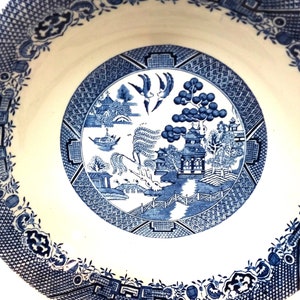 Peut inclure: Un bol en céramique blanche avec un motif de saule bleu et blanc. Le bol présente une scène de paysage chinois traditionnelle avec une pagode, un saule pleureur, un pont et des oiseaux en vol.