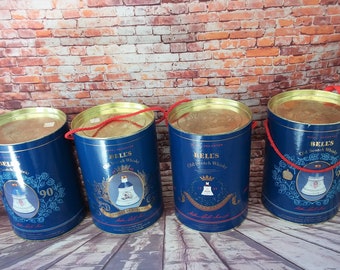 Puede incluir: Cuatro latas azules y doradas con asas de cuerda roja. Cada lata tiene una etiqueta que dice "Bell's Old Scotch Whisky" y muestra una imagen de una campana. Las latas están dispuestas en fila sobre una superficie de ladrillo.