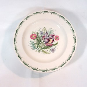 May include: A white ceramic plate with a green border and a floral design featuring a pink and purple tulip, pink and white flowers, and green leaves.