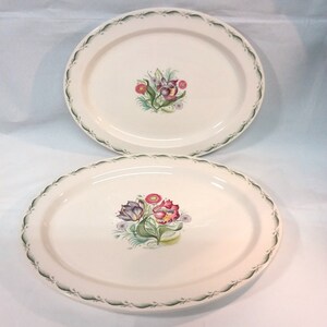 May include: Two white oval ceramic plates with green trim and floral designs. The plates feature a cluster of pink, purple, and white flowers with green leaves.