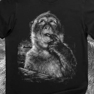 May include: Black t-shirt with a monochrome illustration of a monkey, its finger raised to its mouth, gazing upwards. The design has a detailed, artistic style, with a dark background.