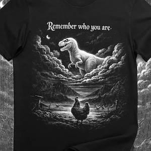 Puede incluir: Camiseta negra con un gráfico de un T-Rex en un cielo nublado sobre una gallina en un campo. El texto "Remember who you are" está encima del dinosaurio, en un diseño monocromático.