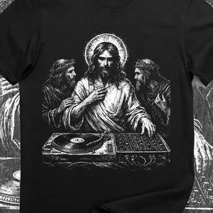 May include: A black short-sleeved t-shirt featuring a white graphic illustration of a central figure with a halo, flanked by two bearded figures, all positioned behind a DJ turntable and mixer setup, rendered in a detailed, sketch-like style.