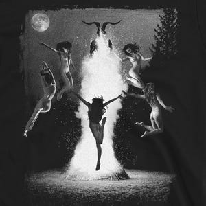 Puede incluir: Camiseta negra con un gráfico en escala de grises que representa varias figuras bailando alrededor de una fuente de luz brillante. El diseño incluye una luna llena, un árbol y un par de cuernos sobre la luz. El tema general es místico y artístico.
