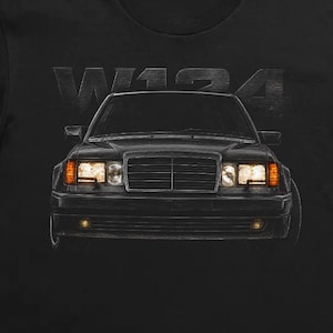 Può includere: Una t-shirt nera a maniche corte con una stampa grafica di un'auto vista frontalmente con i fari accesi. Il testo W124 è stampato in un carattere grigio scuro sopra l'auto. Lo sfondo della maglietta presenta un motivo sbiadito di una calandra.