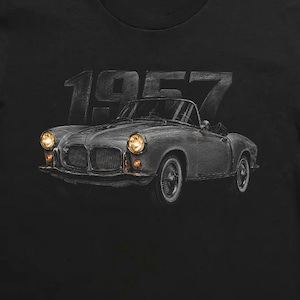 May include: A black short-sleeved t-shirt featuring a graphic of a vintage convertible car in shades of grey with glowing yellow headlights. Above the car, the text 1957 is printed in a bold, dark grey font. The design is centred on the chest.