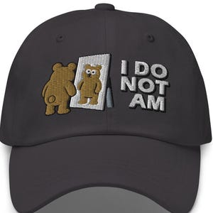 May include: A dark gray baseball cap featuring an embroidered graphic of a brown bear looking into a mirror, with the text I DO NOT AM stitched in white letters to the right of the mirror. The cap has a curved brim and a structured crown.
