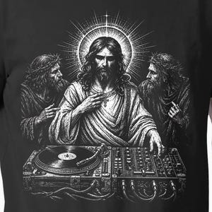 May include: A black t-shirt featuring a white graphic illustration of a central figure with a halo, flanked by two bearded figures, all positioned behind a DJ turntable and mixer setup. The central figure has long hair and wears robes.