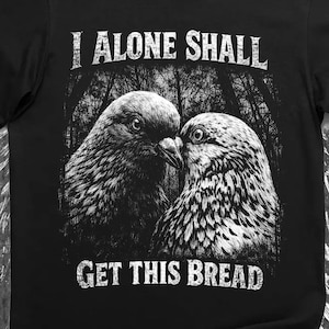 Op de afbeelding: Zwart t-shirt met een afbeelding van twee vogels tegen een bosachtergrond. De tekst "I ALONE SHALL" staat boven de vogels en "GET THIS BREAD" eronder. Het ontwerp is in wit.