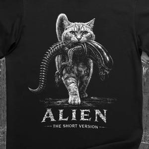 Pode incluir: T-shirt preto com uma ilustração em tons de cinza de um gato carregando uma criatura Alien. O texto "ALIEN - THE SHORT VERSION" está impresso abaixo da imagem. A camisa é feita de um material macio.