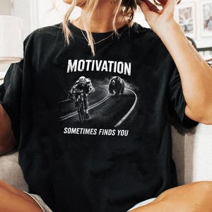 K&ouml;nnte beinhalten: Schwarzes T-Shirt mit dem Wort "MOTIVATION" &uuml;ber einer Grafik eines Radfahrers, der von einem B&auml;ren auf einer kurvenreichen Stra&szlig;e verfolgt wird. Der Text "SOMETIMES FINDS YOU" steht darunter. Ein l&auml;ssiges Kleidungsst&uuml;ck.