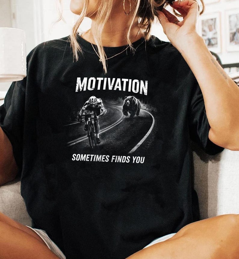 K&ouml;nnte beinhalten: Schwarzes T-Shirt mit dem Wort "MOTIVATION" &uuml;ber einer Grafik eines Radfahrers, der von einem B&auml;ren auf einer kurvenreichen Stra&szlig;e verfolgt wird. Der Text "SOMETIMES FINDS YOU" steht darunter. Ein l&auml;ssiges Kleidungsst&uuml;ck.