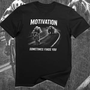 K&ouml;nnte beinhalten: Schwarzes T-Shirt mit dem Wort "MOTIVATION" &uuml;ber einer Grafik eines Radfahrers, der von einem B&auml;ren auf einer Stra&szlig;e verfolgt wird. Der Text "SOMETIMES FINDS YOU" steht darunter. Das T-Shirt hat einen Rundhalsausschnitt.
