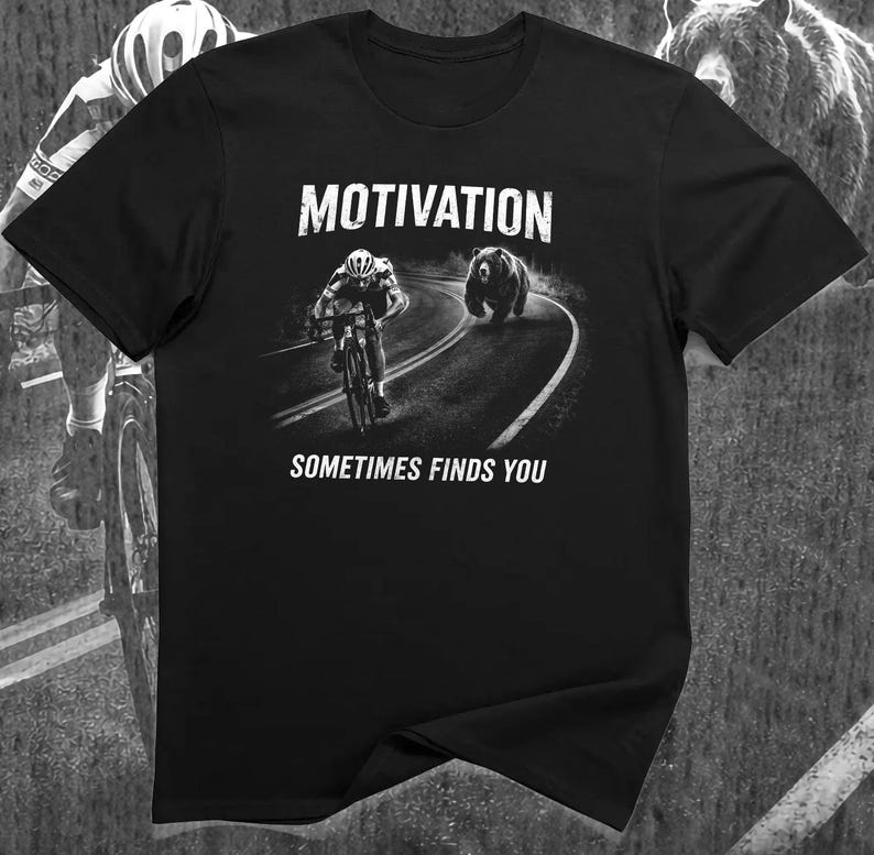 K&ouml;nnte beinhalten: Schwarzes T-Shirt mit dem Wort "MOTIVATION" &uuml;ber einer Grafik eines Radfahrers, der von einem B&auml;ren auf einer Stra&szlig;e verfolgt wird. Der Text "SOMETIMES FINDS YOU" steht darunter. Das T-Shirt hat einen Rundhalsausschnitt.