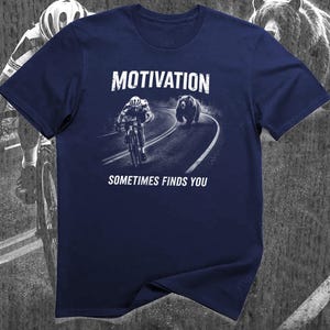 K&ouml;nnte beinhalten: Marineblaues T-Shirt mit einem Aufdruck, der einen Radfahrer zeigt, der von einem B&auml;ren auf einer Stra&szlig;e verfolgt wird. Die Worte "MOTIVATION" und "SOMETIMES FINDS YOU" sind aufgedruckt.