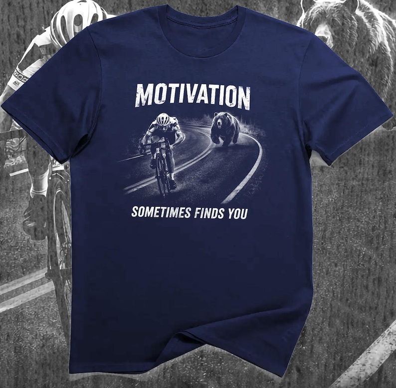 K&ouml;nnte beinhalten: Marineblaues T-Shirt mit einem Aufdruck, der einen Radfahrer zeigt, der von einem B&auml;ren auf einer Stra&szlig;e verfolgt wird. Die Worte "MOTIVATION" und "SOMETIMES FINDS YOU" sind aufgedruckt.