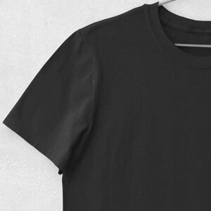 K&ouml;nnte beinhalten: Ein einfarbiges schwarzes T-Shirt mit kurzen &Auml;rmeln. Das Kleidungsst&uuml;ck h&auml;ngt an einem Metallb&uuml;gel vor einer hellen Wand. Der Stoff scheint weich zu sein.