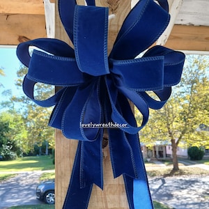 May include: A large, navy blue velvet bow with long streamers is attached to a wooden post. The bow is made of wide, luxurious ribbon and is displayed outdoors. The text "lovelywreathdecor" is visible on the top right.