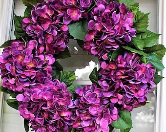 May include: A purple hydrangea wreath with green leaves. The wreath is made of artificial flowers and is ideal for decorating a door or wall.