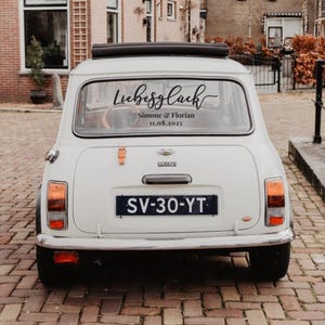 May include: A white Mini car viewed from the rear, featuring a black decal on the back window with the script text "Liebesgl&uuml;ck" above the names "Simone & Florian" and the date "11.08.2025". The car has a black license plate reading "SV-30-YT".