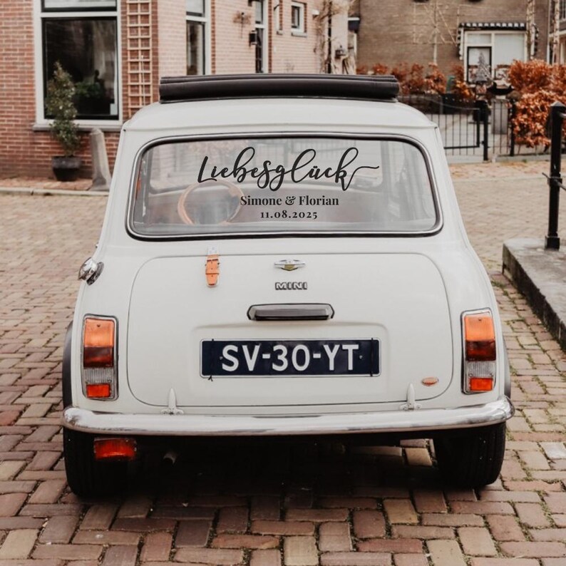 May include: A white Mini car viewed from the rear, featuring a black decal on the back window with the script text "Liebesgl&uuml;ck" above the names "Simone & Florian" and the date "11.08.2025". The car has a black license plate reading "SV-30-YT".