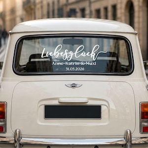 May include: A white vinyl decal applied to the rear window of a cream-colored vintage car. The decal features the word Liebesgl&uuml;ck in a cursive script, followed by the names Anne-Katrin & Maxi and the date 31.03.2026 in a clean font.