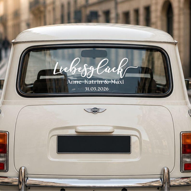 May include: A white vinyl decal applied to the rear window of a cream-colored vintage car. The decal features the word Liebesgl&uuml;ck in a cursive script, followed by the names Anne-Katrin & Maxi and the date 31.03.2026 in a clean font.