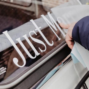 May include: A white vinyl decal with the text Just Married in a serif font being applied to a dark glass surface. The lettering is cut out in a clean, elegant style, showing the individual letters being pressed onto the window of a vehicle.