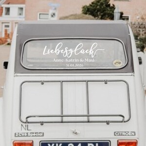 May include: A white vinyl decal applied to the rear window of a white Citroen 2CV car. The decal features the script text Liebesgl&uuml;ck, followed by the names Anne-Katrin & Maxi and the date 31.03.2026 in a clean, sans-serif font style.