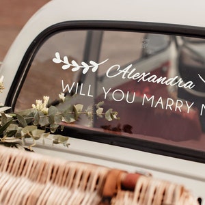 May include: A white car with a floral wreath and the text "Alexandra Will You Marry Me?" written on the back window.