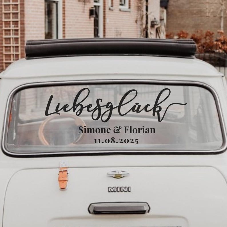May include: A white car rear window featuring a black vinyl decal with the word Liebesgl&uuml;ck in a cursive script font, followed by the names Simone & Florian and the date 11.08.2025 in a clean, sans-serif typeface centered on the glass.