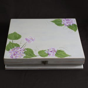 May include: A white wooden box with a hinged lid, hand-painted with purple hydrangea flowers and green leaves. The box has a metal clasp and is likely used for storage or display. The floral design adds a touch of elegance.