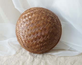 May include: A circular basket made from woven brown material, featuring a geometric pattern. The basket has a domed shape and is set against a light-coloured backdrop. The basket's diameter is approximately 25 cm.