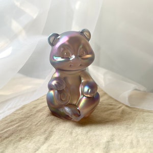 May include: A small iridescent glass bear figurine with a pink hue. The bear is sitting with its paws crossed and has a round, button-like nose.
