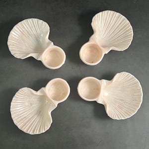 May include: Four seashell-shaped dishes with a small circular bowl attached. The dishes are off-white with subtle pink and peach tones, and feature a ribbed texture. The bowls are also off-white with a pinkish hue.