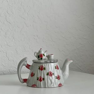 Peut inclure: Une théière en céramique blanche avec un motif floral. La théière a un petit couvercle avec une tasse à thé assortie sur le dessus. La théière est décorée de roses roses et de feuilles vertes.