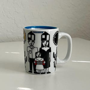 May include: White ceramic mug with a blue rim. The mug features a black and white illustration of a family with a cactus and a sun. The family is wearing black and white clothing. The woman is wearing a black dress and the man is wearing a white shirt. The child is holding a red accordion.
