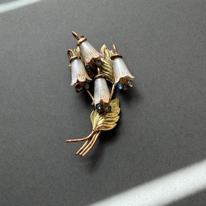 Può includere: Spilla floreale vintage con fiori a forma di campana in bianco e oro. La spilla presenta foglie e steli dorati, con piccole pietre preziose blu. Un gioiello classico, lungo circa 5 cm.