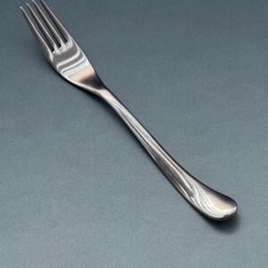 Könnte beinhalten: Eine Edelstahlgabel mit vier Zinken und einem gebogenen Griff. Die Gabel besteht aus glänzendem Metall und ist zum Essen bestimmt. Der Griff hat ein dezentes, dekoratives Muster. Es ist ein Besteckteil.